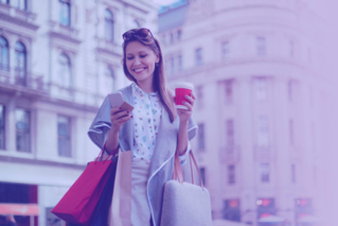Woman smiling while holding a cup of coffee and checking her cell phone with shopping bags in her hand.