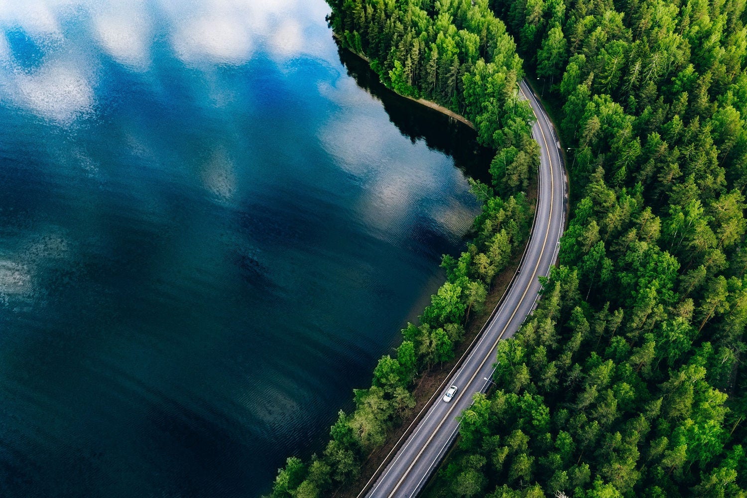Aerial view of the road between the lush green summer forest and the blue lake in Finland