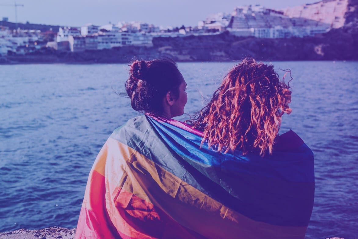 Two people sitting by the sea, covered by a rainbow flag.