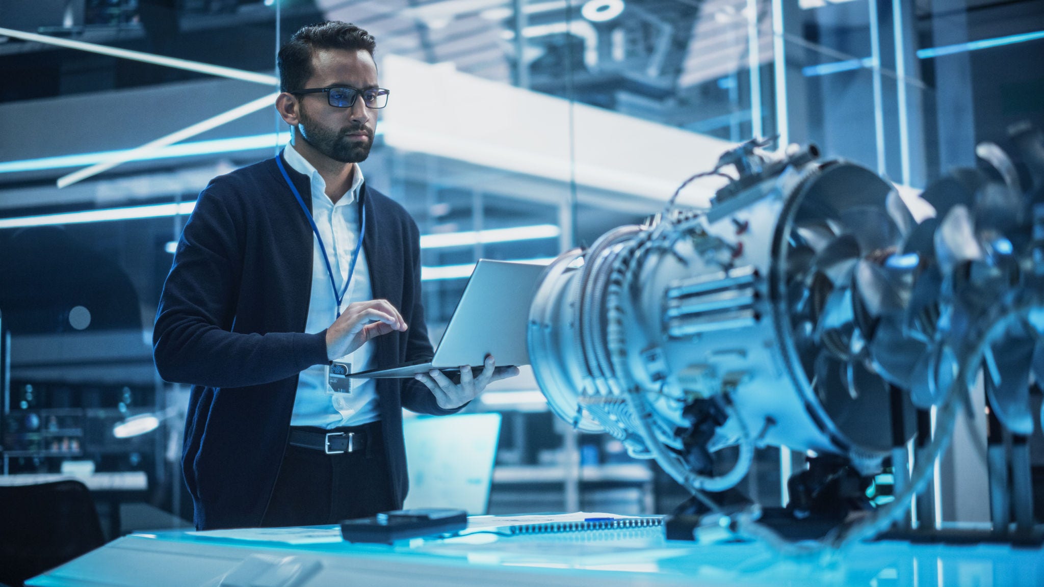 Young Industrial Engineer Working on a Futuristic Jet Engine, Standing with Laptop Computer in Scientific Technology Lab. Scientist Developing a New Electric Motor in a Research Facility. Young Industrial Engineer Working on a Futuristic Jet Engine, Standing with Laptop Computer in Scientific Technology Lab. Scientist Developing a New Electric Motor in a Research Facility.