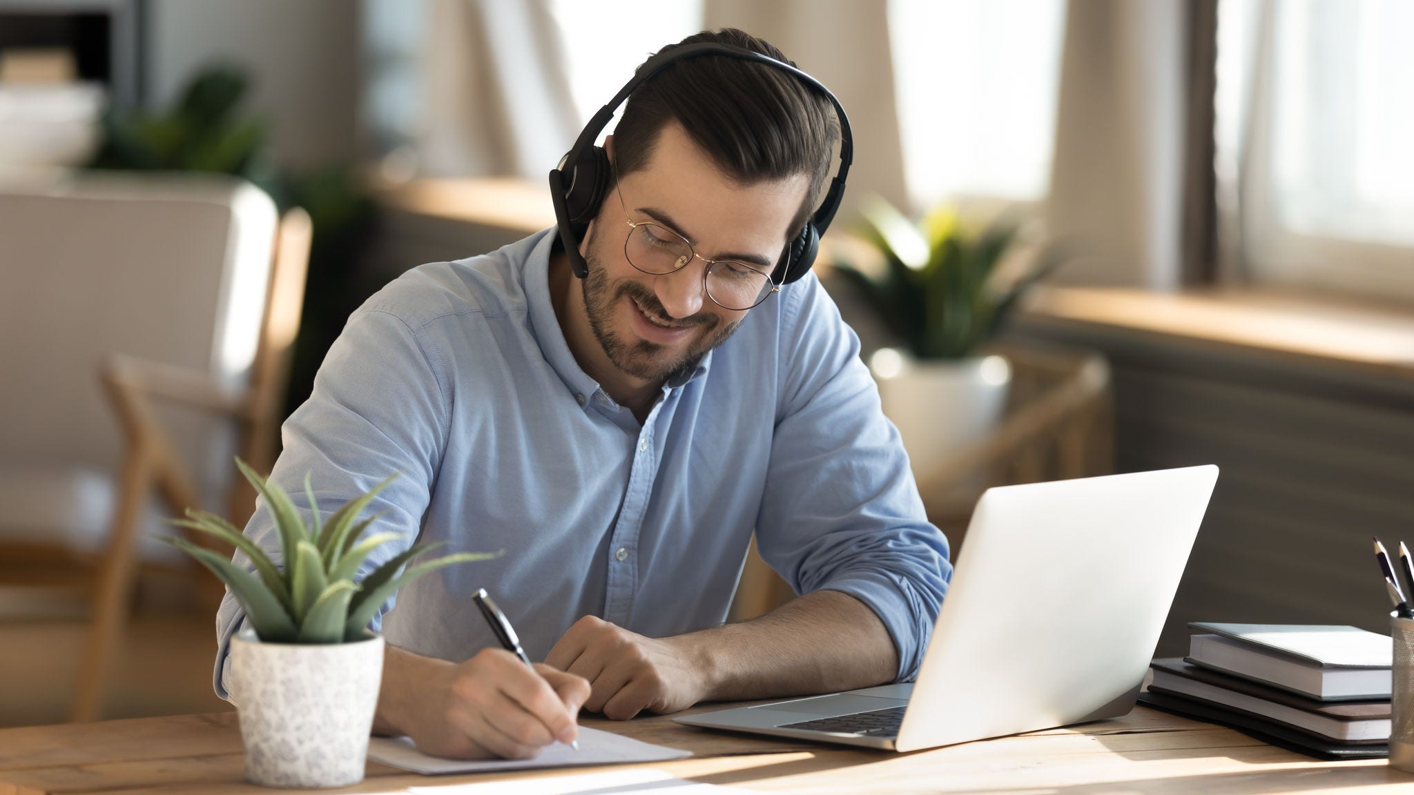 Smiling young Caucasian man in headphones glasses sit at desk work on laptop making notes. Happy millennial male in earphones watch webinar or training course or computer, study online from home.