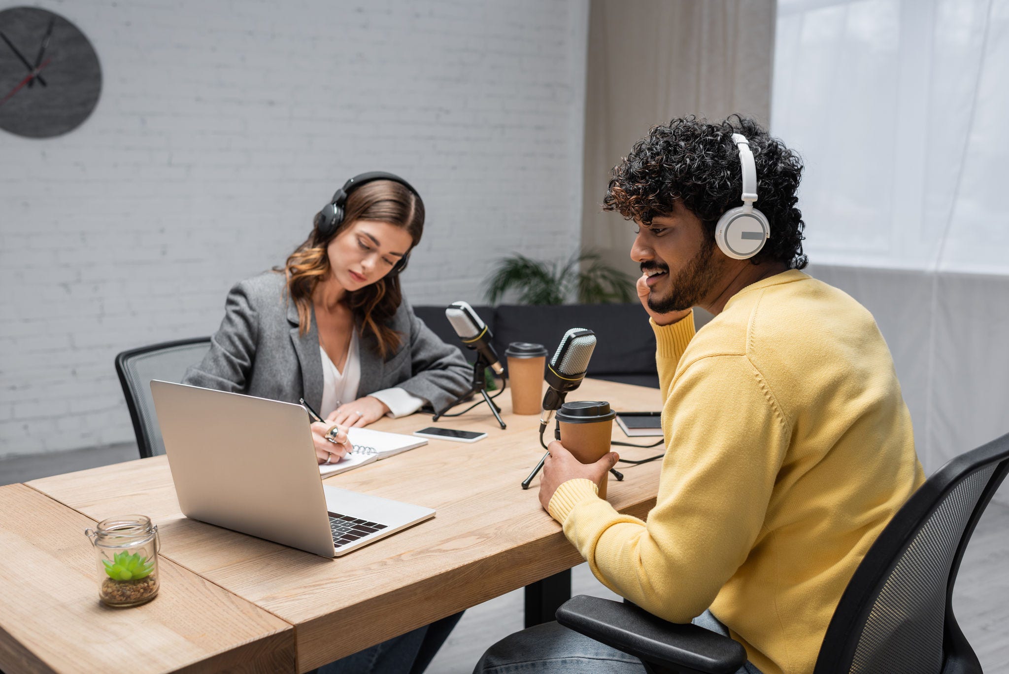 cheerful and curly indian man in headphones and yellow jumper holding coffee to go and looking at laptop near microphones and radio host writing in notebook near smartphone on table in radio studio