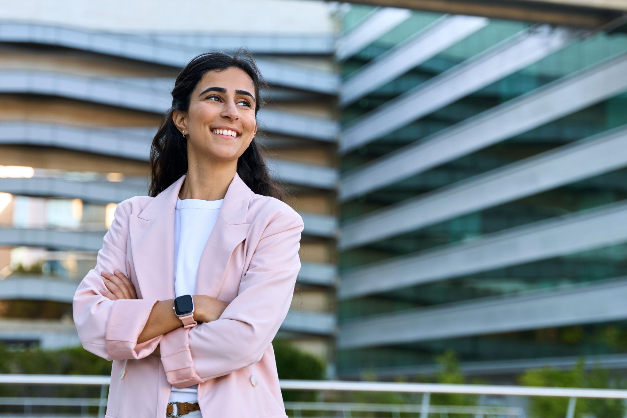 Confident portrait of young middle eastern Israel businesswoman standing with crossed hands at office building. Successful smiling indian or arabic woman in business suit looking aside. Copy space