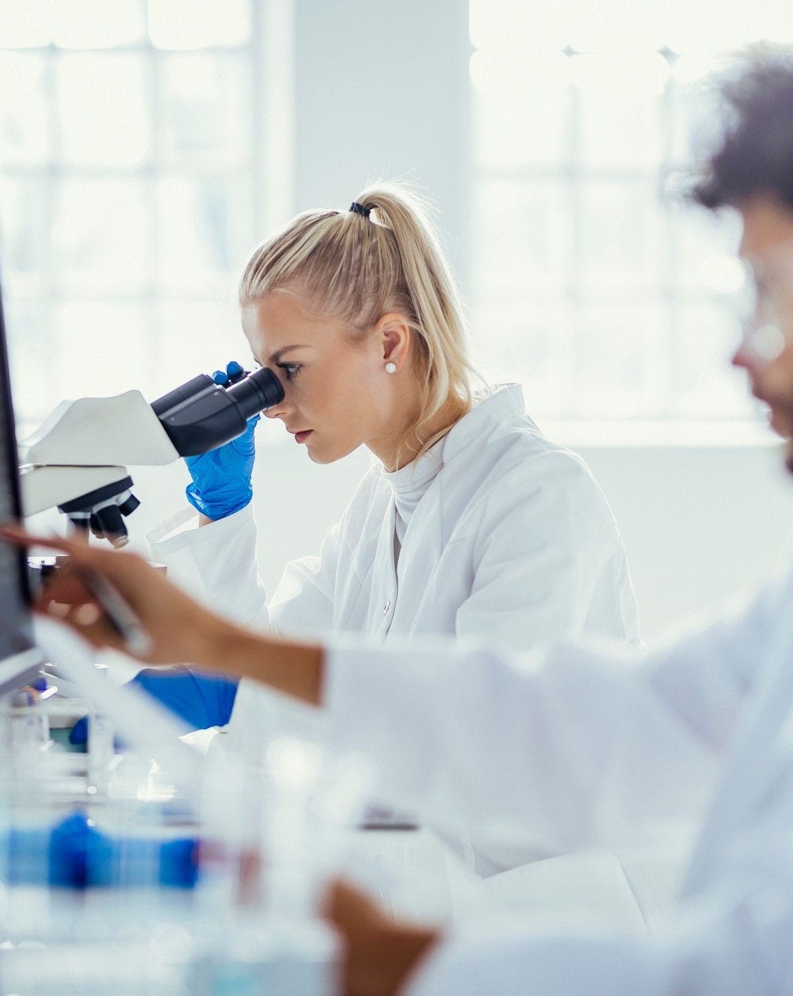 A Middle Eastern male and Caucasian female, clad in white lab coats, concentrate on analyzing specimen under a microscope, amidst scientific tools in a bright laboratory setting.