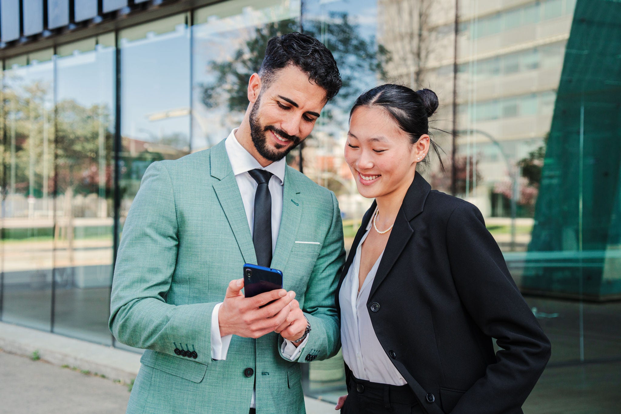 A couple of young businesspeople watching their successful stock shares on smartphone. Two partners smiling using a cellphone to invest on market. Coworkers with a mobile phone at corporate workplace. High quality photo