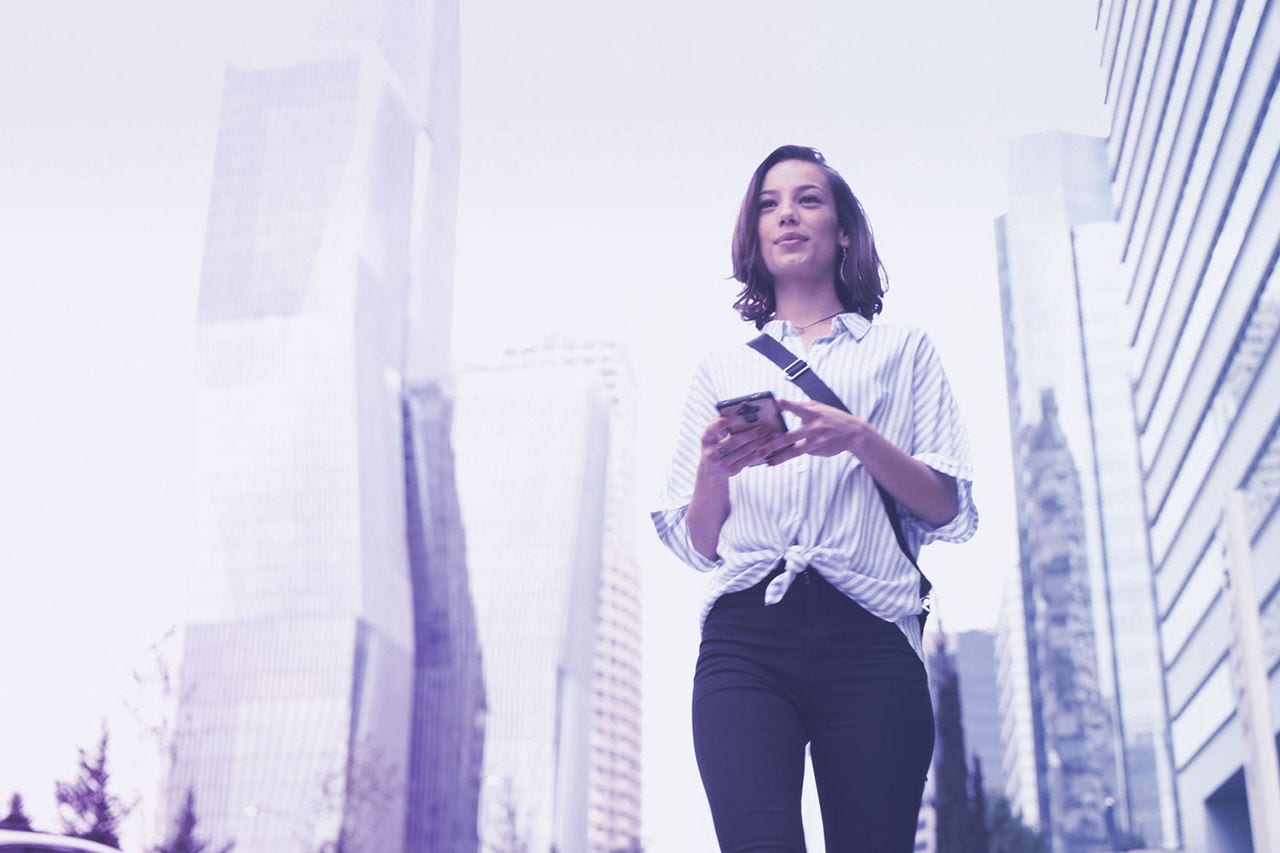 mujer con camisa de rayas verticales, blancas y grises con un teléfono móvil en la mano, su mirada se dirige hacia el móvil mientras camina