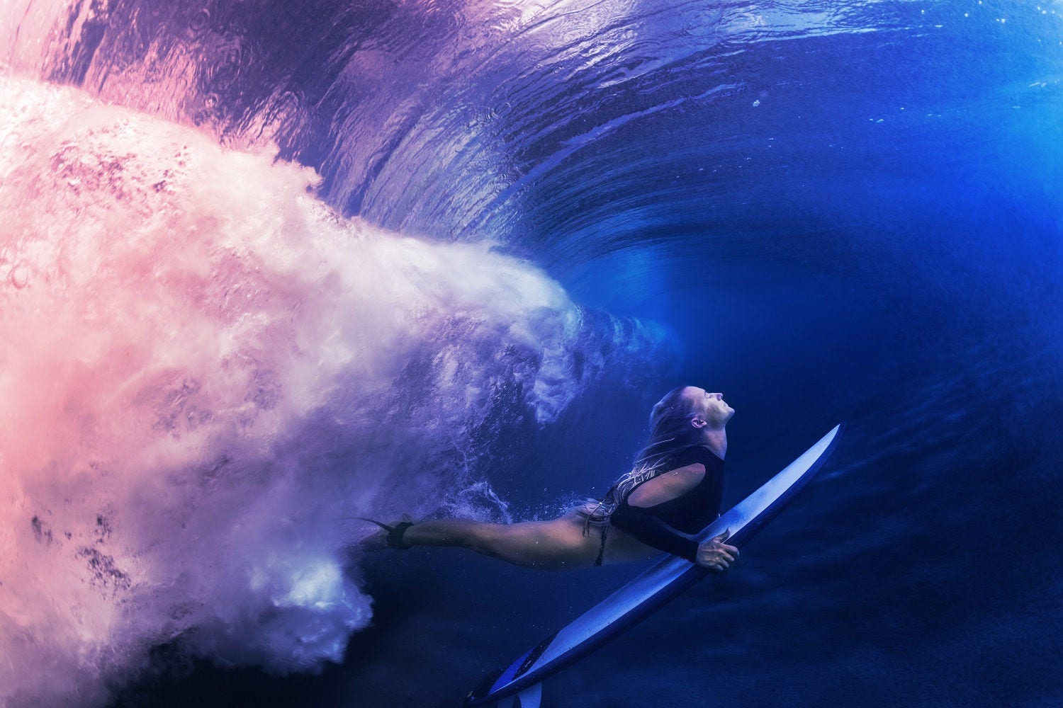 A blonde surfer girl underwater doing duck dive holding surfing board left behind air bubbles in blue water background under big ocean wave