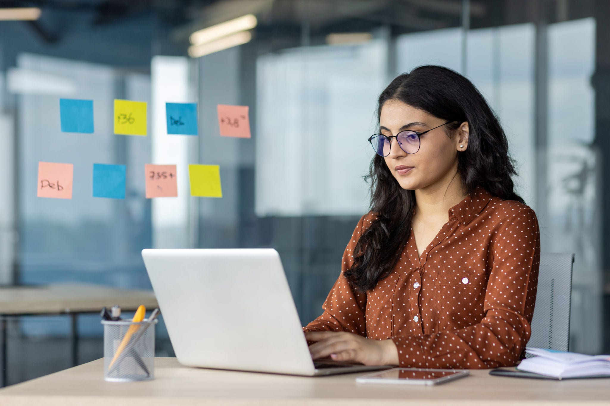 A dedicated office worker is focused on her laptop, surrounded by sticky notes on a glass wall. Her concentration highlights productivity and organization in a modern office setting.