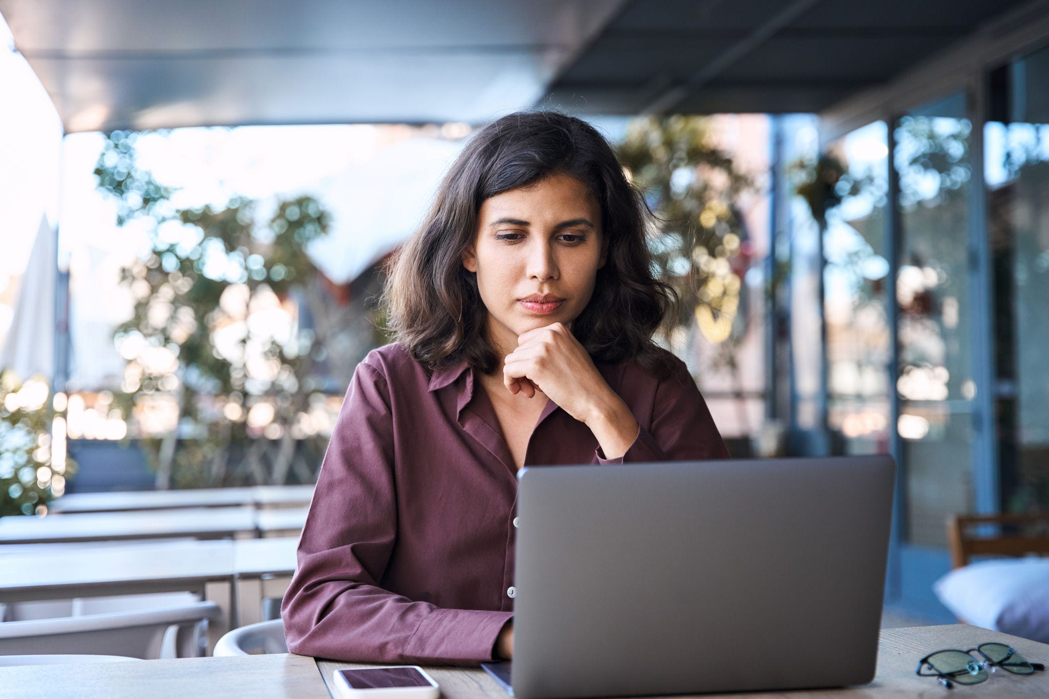 Thoughtful serious freelance entrepreneur business woman working on laptop computer remotely outdoors. Thinking focused busy latin, middle eastern indian young female worker using pc for study, work