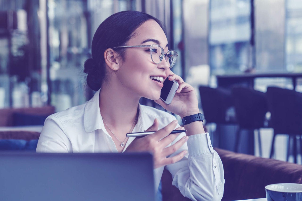 Woman wearing glasses talking on her mobile phone while working on a laptop in a café.