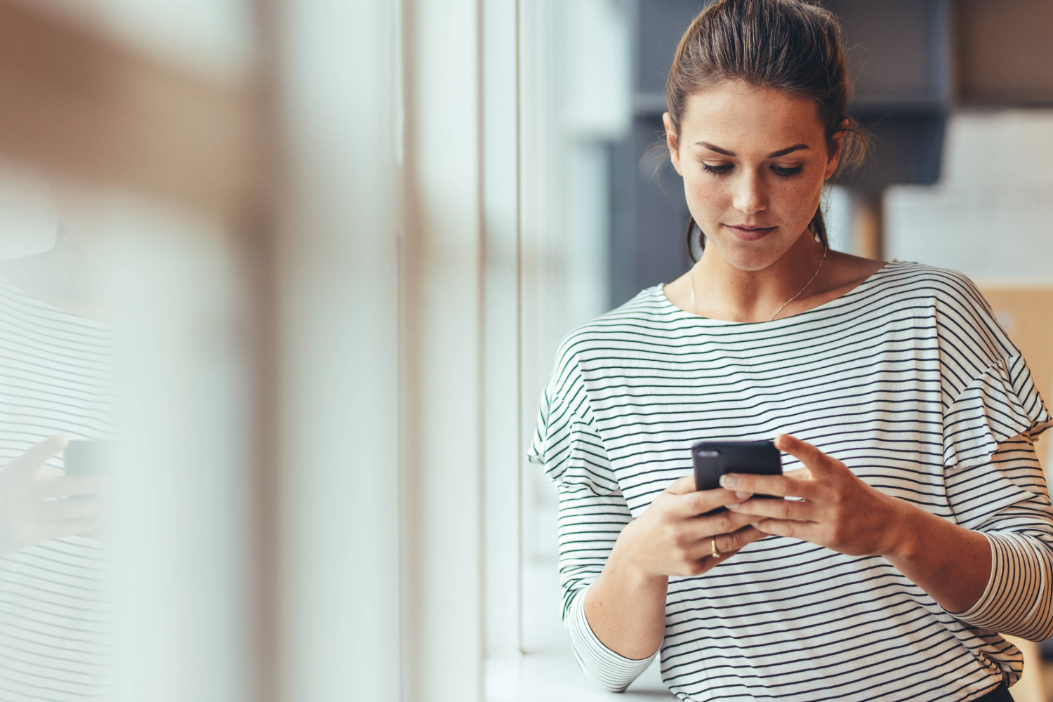 Person standing indoors looking down at a smartphone, with a blurred windowed background