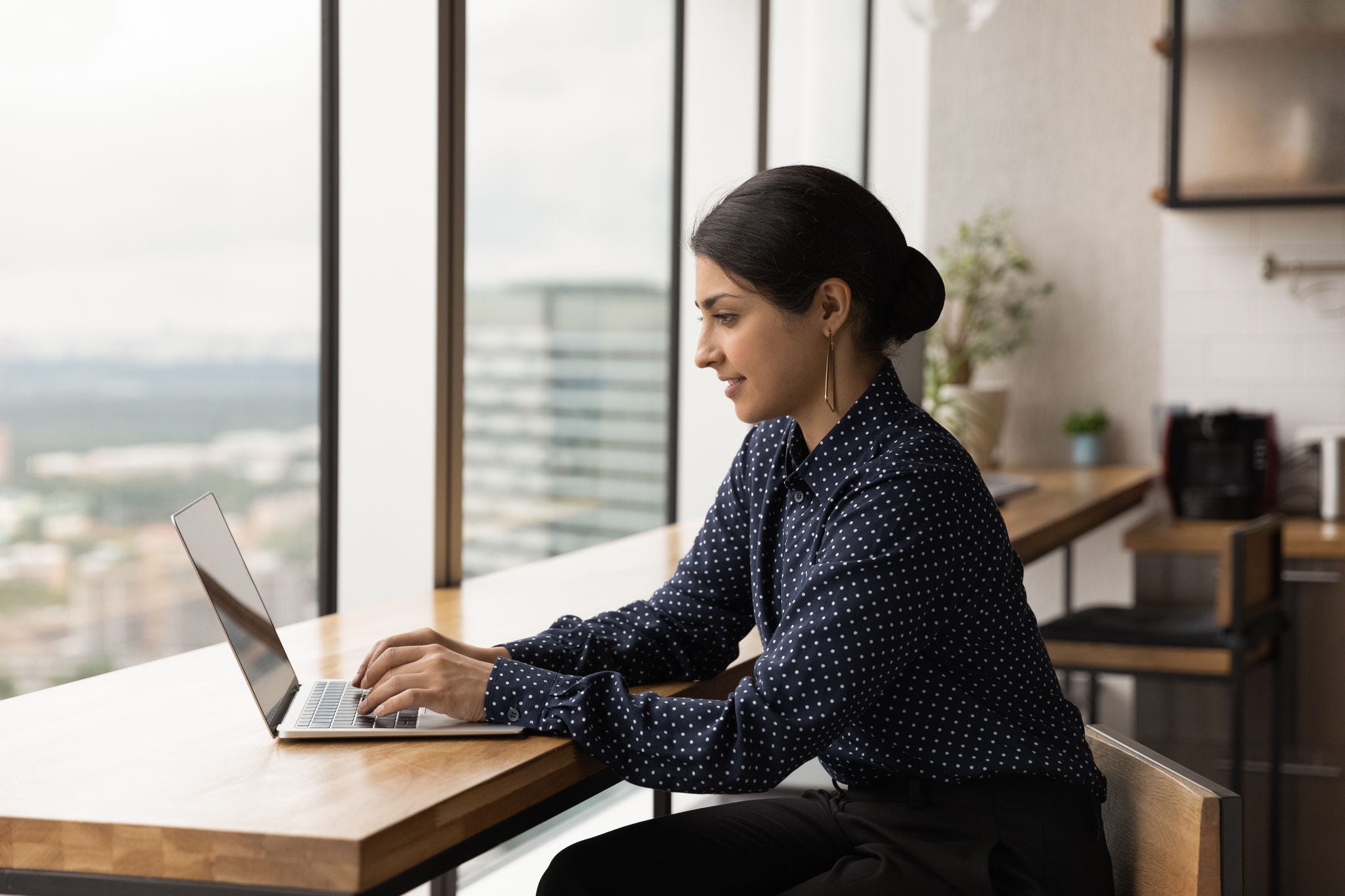 Happy young female indian employee working distantly on online project on computer, typing email communicating with client, preparing report or presentation, sitting at table at modern office room.