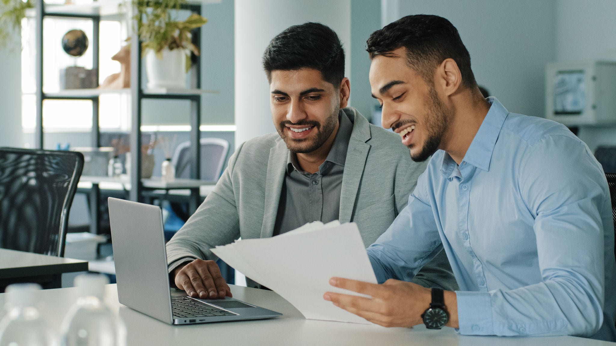 Cheerful positive friendly businessmen partners colleagues holding document contract looking at laptop screen discuss ideas having fun during break in office. Good business relationships, concept. High quality photo