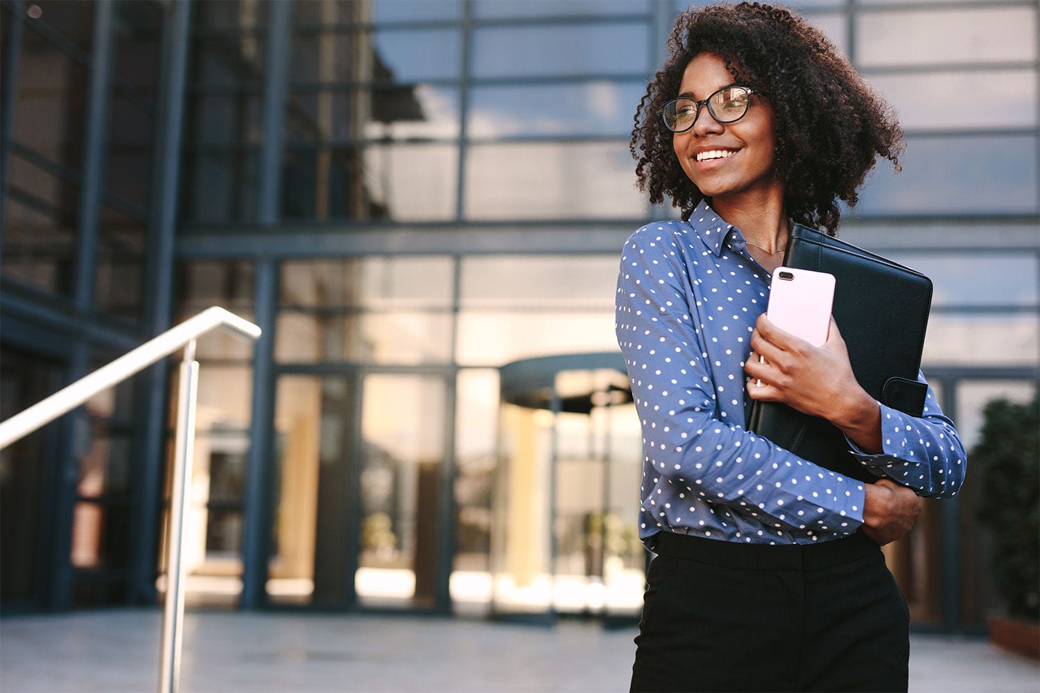 Happy business woman standing outside a office building Woman with mobile phone and file standing outdoors and looking away. Business woman standing outside a office building.
