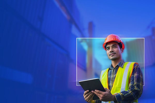 Person wearing an orange safety helmet and a high visibility yellow safety vest stands at an industrial site, holding a tablet.