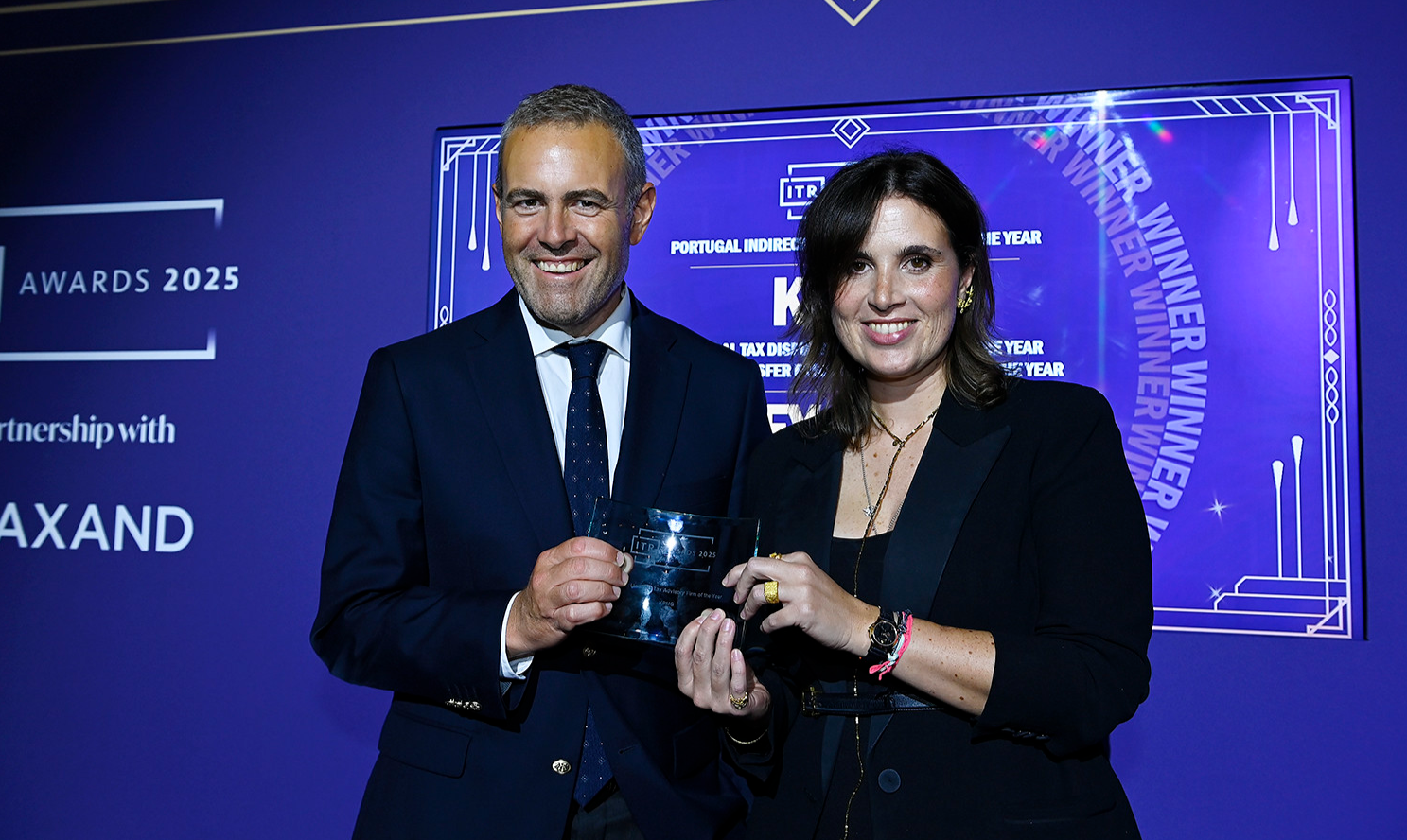 Duas pessoas a sorrir enquanto seguram um prémio durante a cerimónia ITR Awards 2025, diante de um fundo roxo com o logótipo do evento.